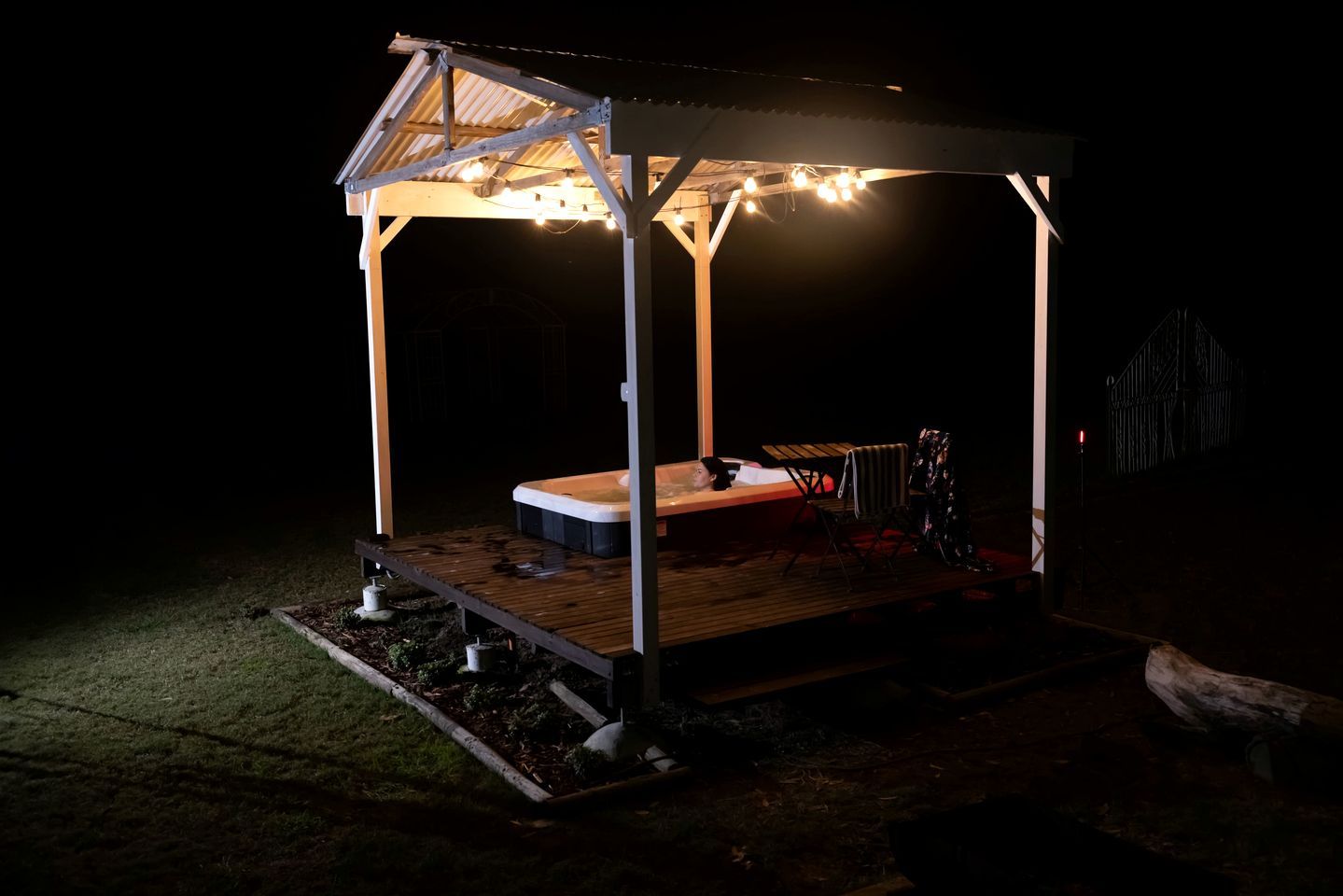 Un espace extérieur abrité avec un jacuzzi, une terrasse en bois, une structure en métal et des guirlandes lumineuses.