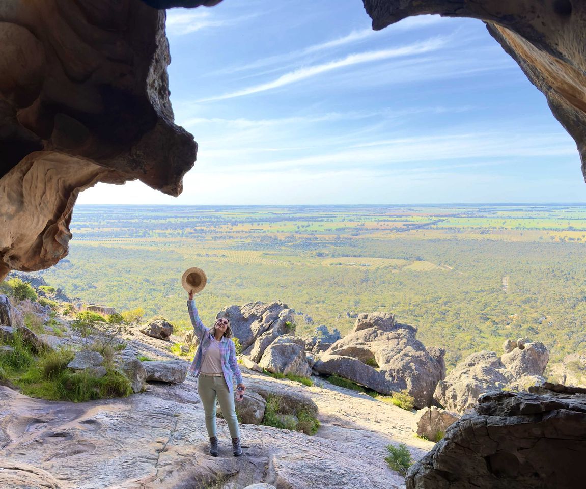 A person standing on a rocky outcrop with a view of a vast landscape through a natural rock arch.