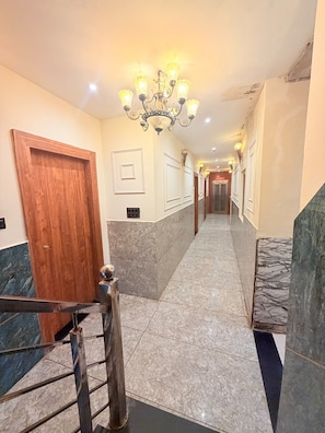 Un pasillo de hotel con puerta de madera, suelo de mármol, barandilla de metal y un candelabro.