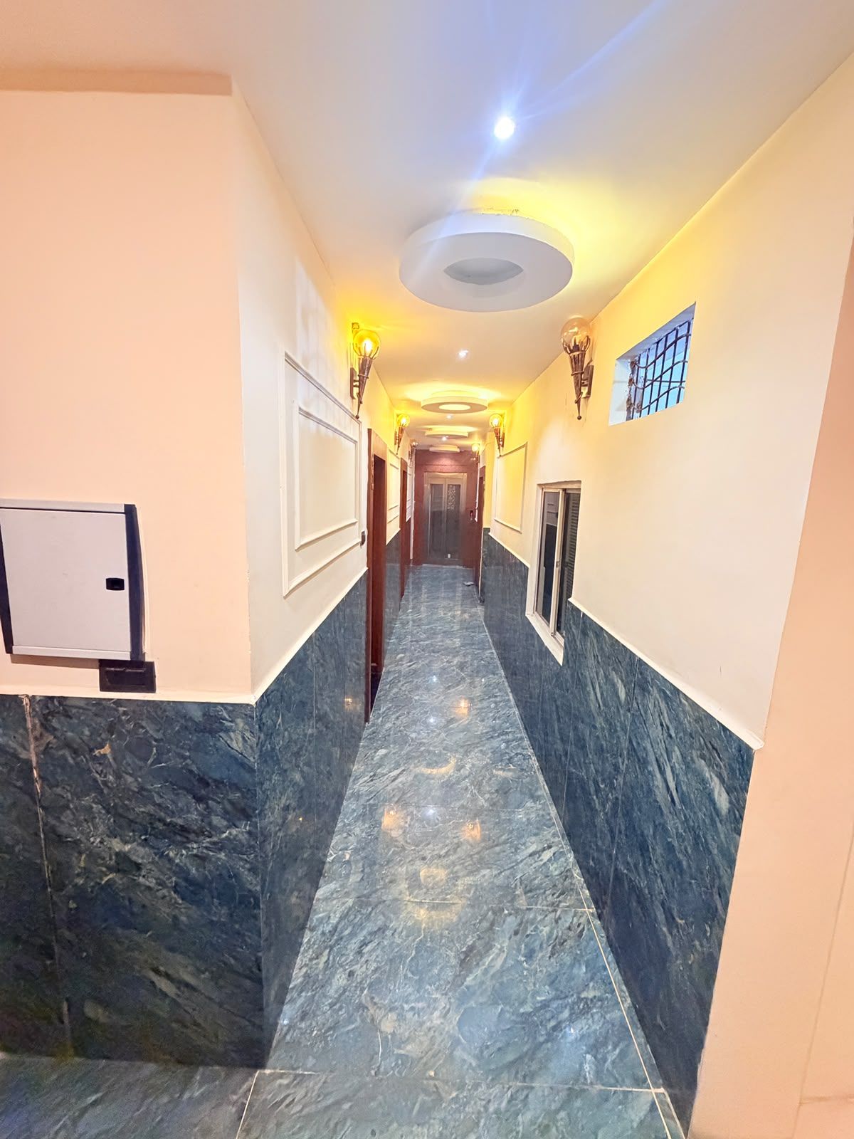 A hallway with marble flooring, wall-mounted light fixtures, and a door at the end.