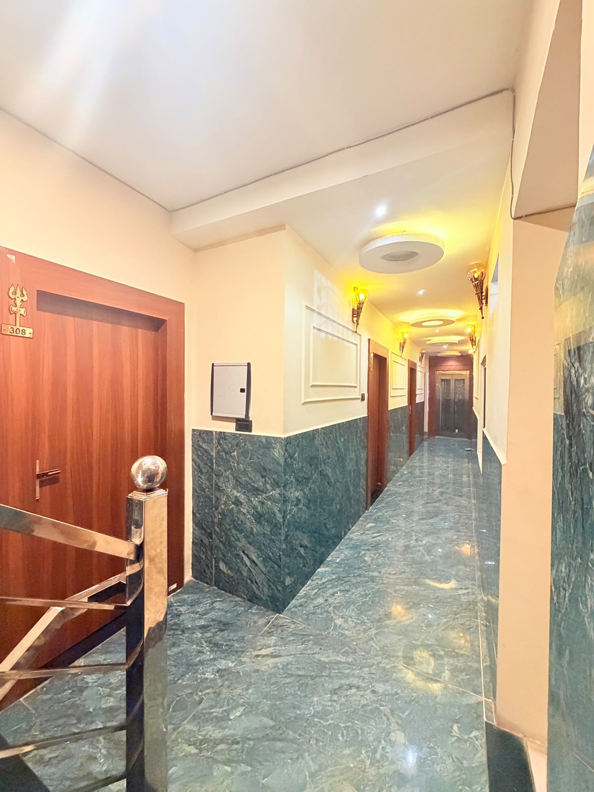 A hotel hallway with marble flooring, a wooden door with a number, and a staircase with a metal railing.