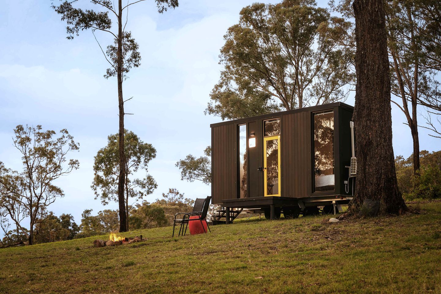 A small, single-story cabin with a door and windows, situated on a grassy slope with trees in the background.