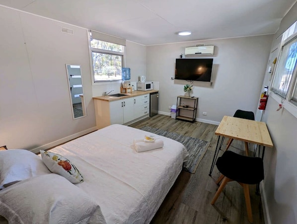 A bedroom with a bed, a desk, a chair, a TV, and a kitchenette.