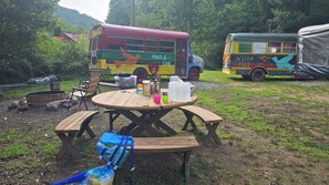 Un montaje para picnic con una mesa y bancos de madera, rodeado de follaje y dos autobuses de colores al fondo.