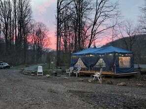 A blue tent with a table and chairs set up on a gravel area, surrounded by trees and a sunset in the background. - C1 Bohemian Bus Retreat | Two Vintage Buses (Parsons)