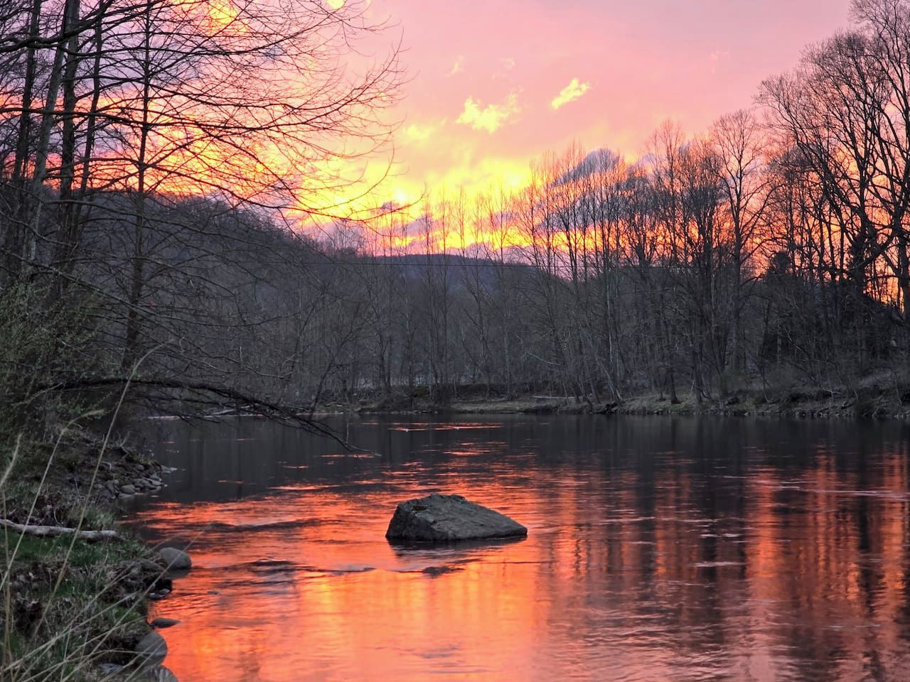 A river with a rock in the middle, surrounded by trees and a sunset sky.