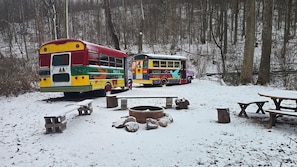 Two colorful buses parked in a snowy forest setting with picnic tables and a fire pit.