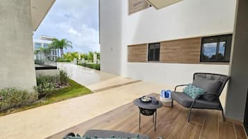 Uma área de estar moderna ao ar livre, com um sofá, cadeiras, uma pequena mesa e vista para um edifício ao fundo.
