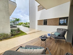 Uma área de estar moderna ao ar livre, com um sofá, cadeiras, uma pequena mesa e vista para um edifício ao fundo.
