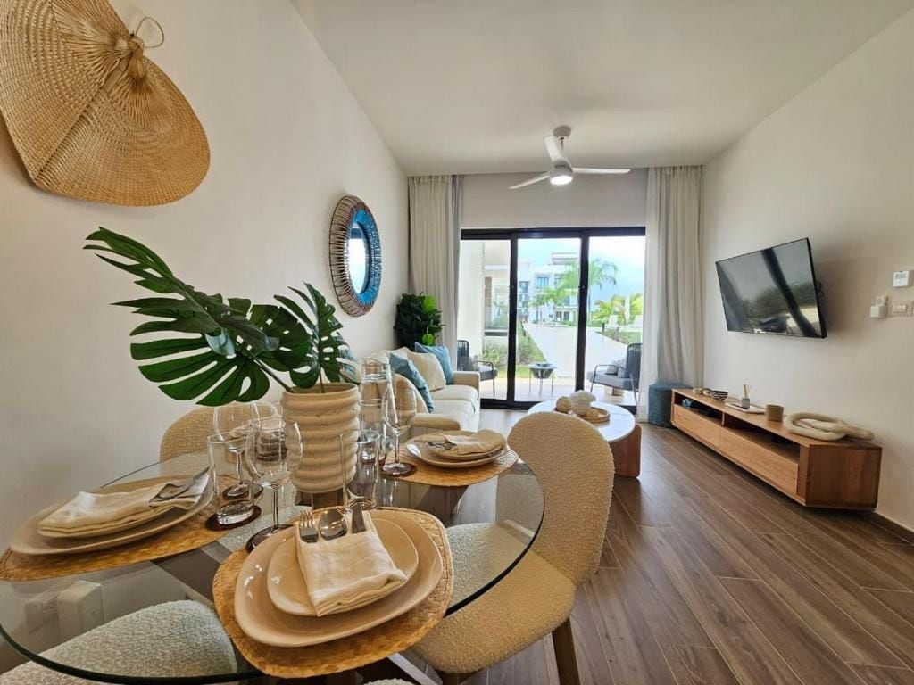 A dining area with a glass table, chairs, and a plant. There is a TV mounted on the wall and a balcony with a view.