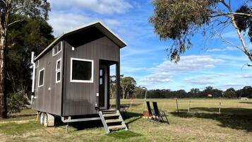 A small, single-story cabin with a door and windows, situated on a grassy area with trees in the background.