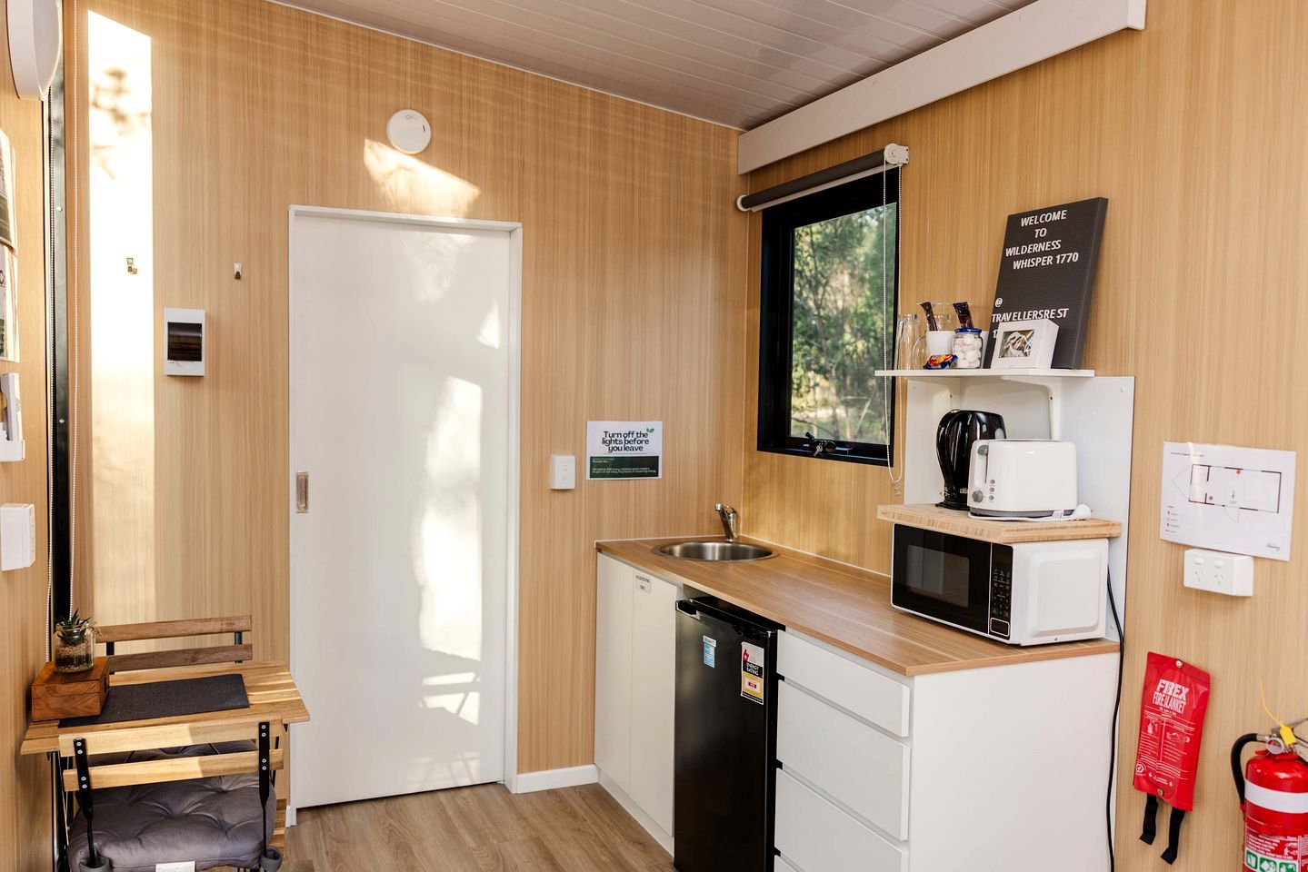 A compact kitchen with a sink, microwave, and a small countertop. There's a door with a sign instructing to turn off the lights before leaving. A fire extinguisher is mounted on the wall.