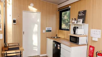 A compact kitchen with a sink, microwave, and a small countertop. There's a door with a sign instructing to turn off the lights before leaving. A fire extinguisher is mounted on the wall.