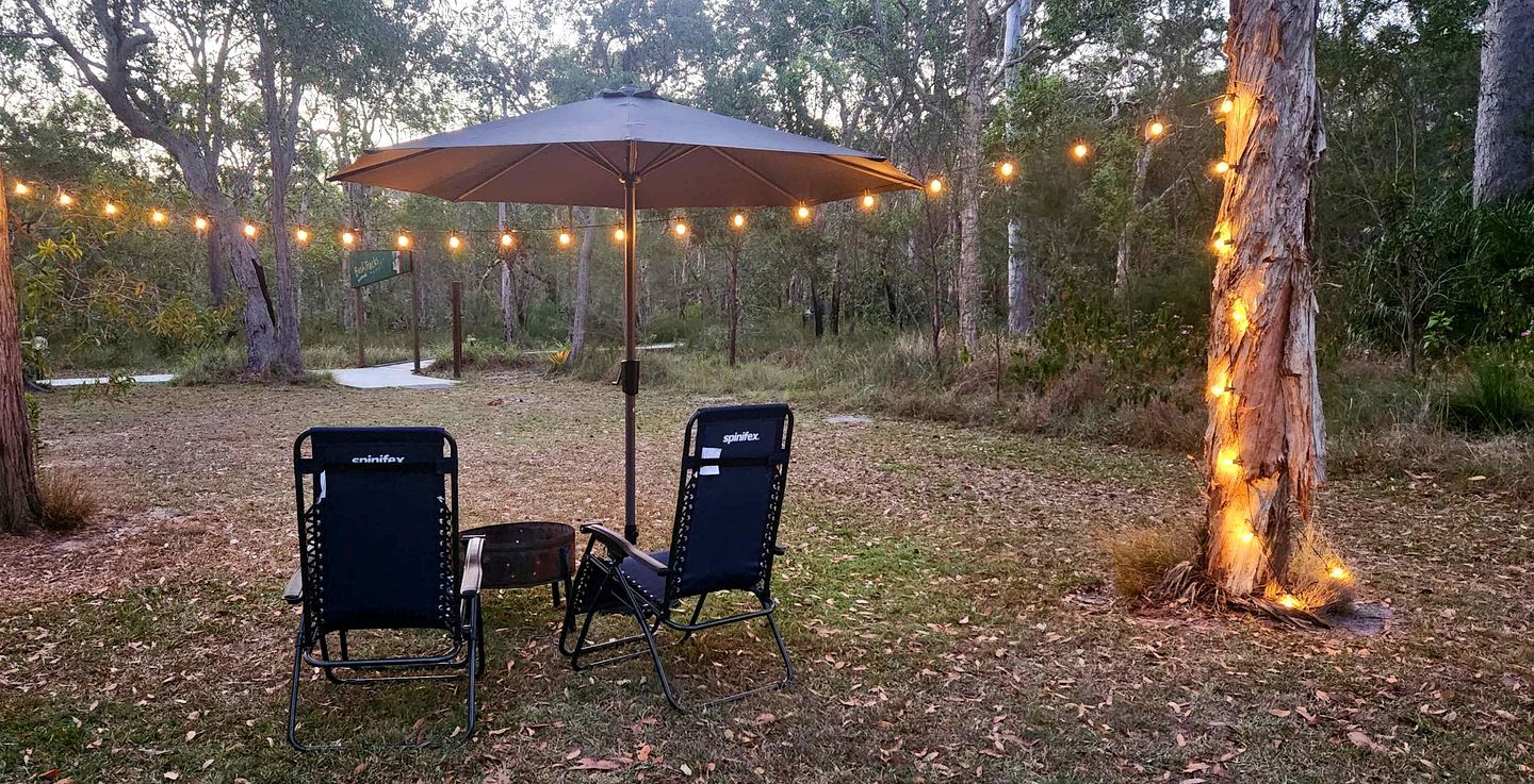 Un espacio de estar al aire libre con dos sillas y una mesa bajo una sombrilla, rodeado de árboles y luces colgantes.