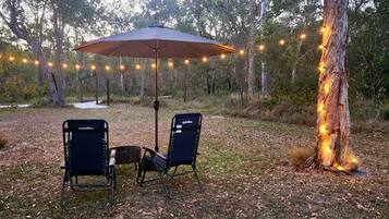 Un espacio de estar al aire libre con dos sillas y una mesa bajo una sombrilla, rodeado de árboles y luces colgantes.