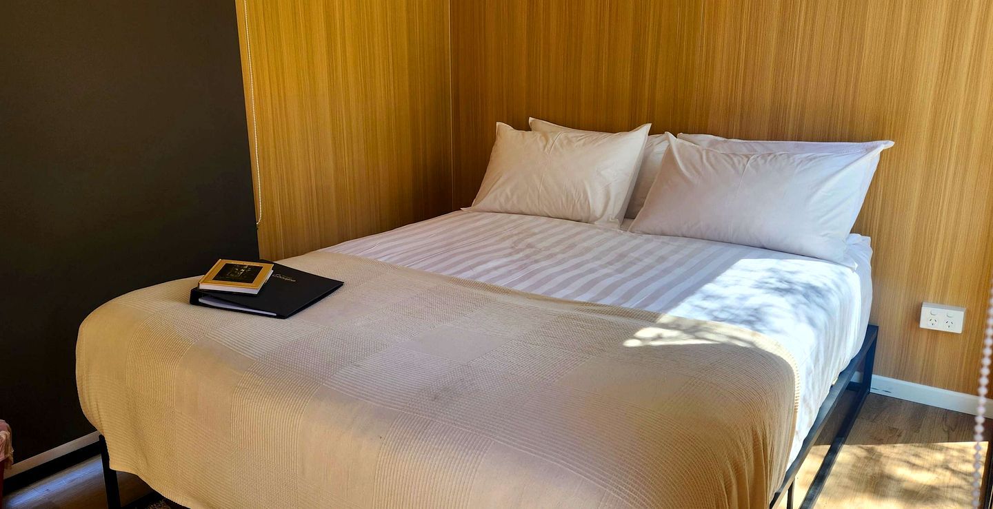 Un lit bien fait, avec un livre posé sur la table de chevet, une tête de lit en bois et une prise électrique sur le mur.