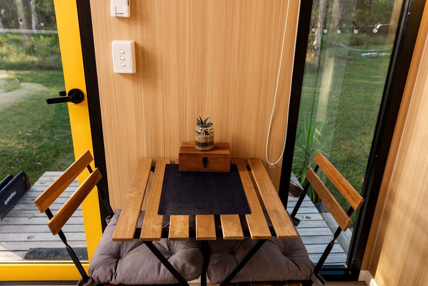 A small wooden table with a black mat, two wooden chairs, a small wooden box, and a potted plant on a balcony with a glass door.