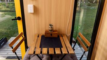 A small wooden table with a black mat, two wooden chairs, a small wooden box, and a potted plant on a balcony with a glass door.