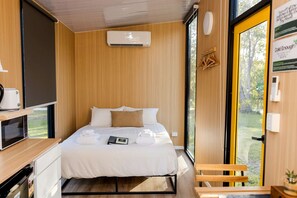 A modern bedroom with a bed, a bedside table, a microwave, and a window with a view of greenery.