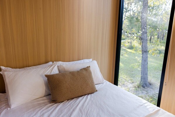 A bed with white linens and a single brown pillow, set against a wooden wall with a window overlooking a natural landscape.