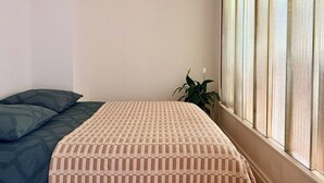 Ein Zimmer mit einem Bett, einem Fenster mit Jalousien und einer Zimmerpflanze.