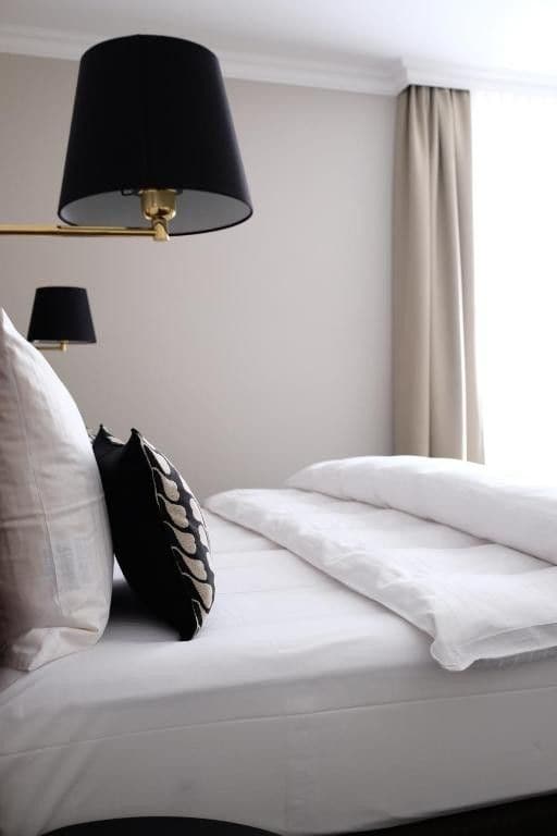 A neatly made bed with white linens and a black pillow, a black lamp, and a beige curtain.