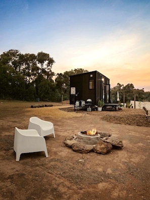 A modern outdoor seating area with a fire pit, a dark-colored building, and trees in the background.