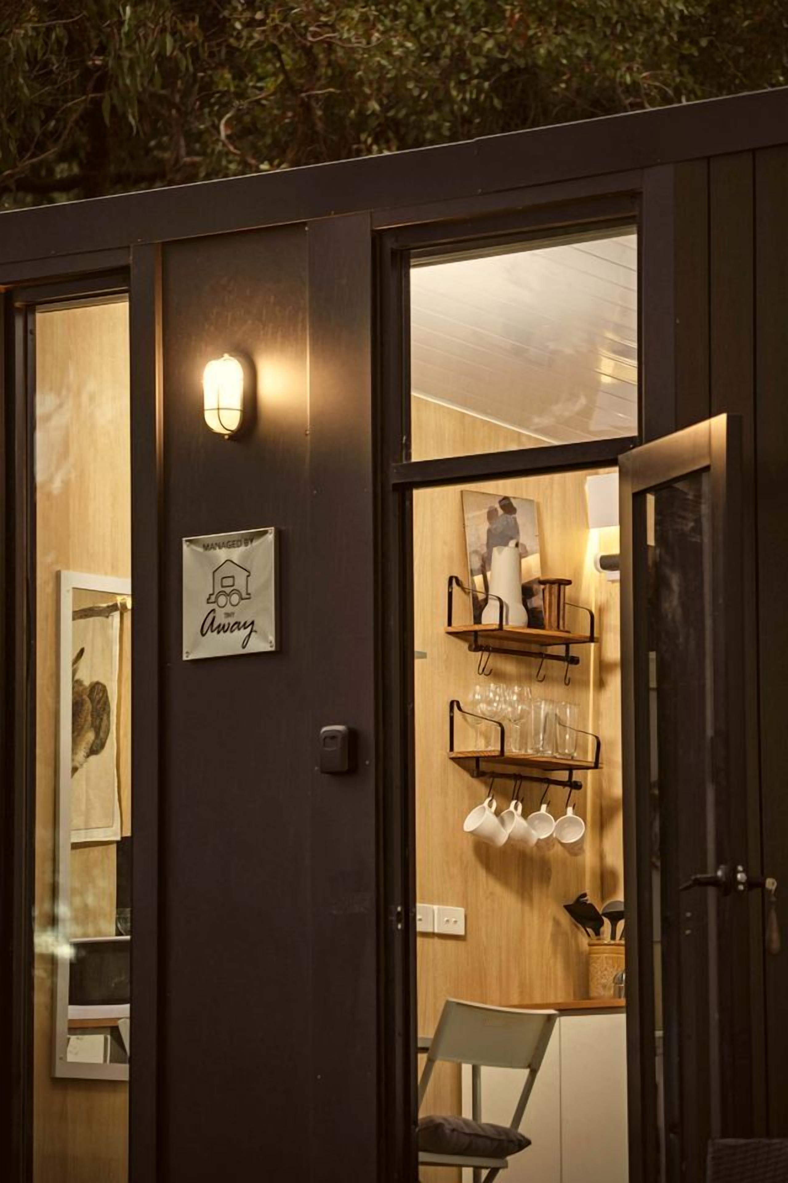 A room with a wooden door, a wall-mounted light, a sign, and a shelf with cups and a vase.