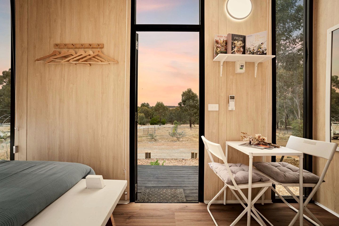 A modern bedroom with a large window, a bed, a desk with a chair, and a small shelf with books.