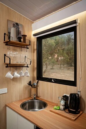 A compact kitchen with a built-in sink, a window with a view of trees, and a shelf with cups and glasses.