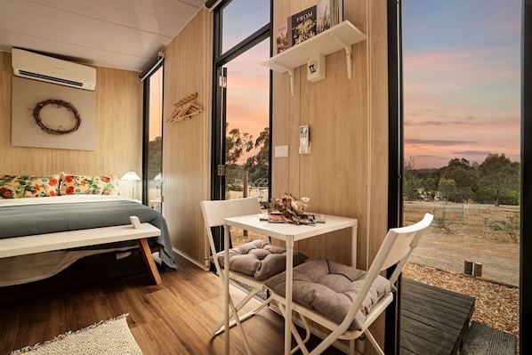 A modern bedroom with a bed, a small table, and a chair. There is a large window providing a view of the outdoors.