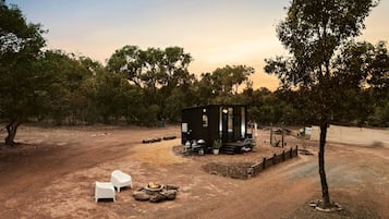 Une petite cabane avec une véranda, entourée d’arbres et bordée d’un sol en terre battue.