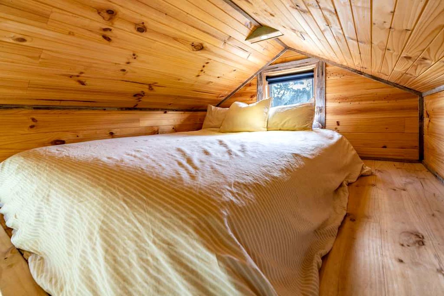 A cozy wooden cabin room with a sloped ceiling, a single bed with a white bedspread, and a window providing natural light.