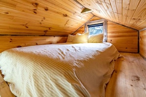 A cozy wooden cabin room with a sloped ceiling, a single bed with a white bedspread, and a window providing natural light.