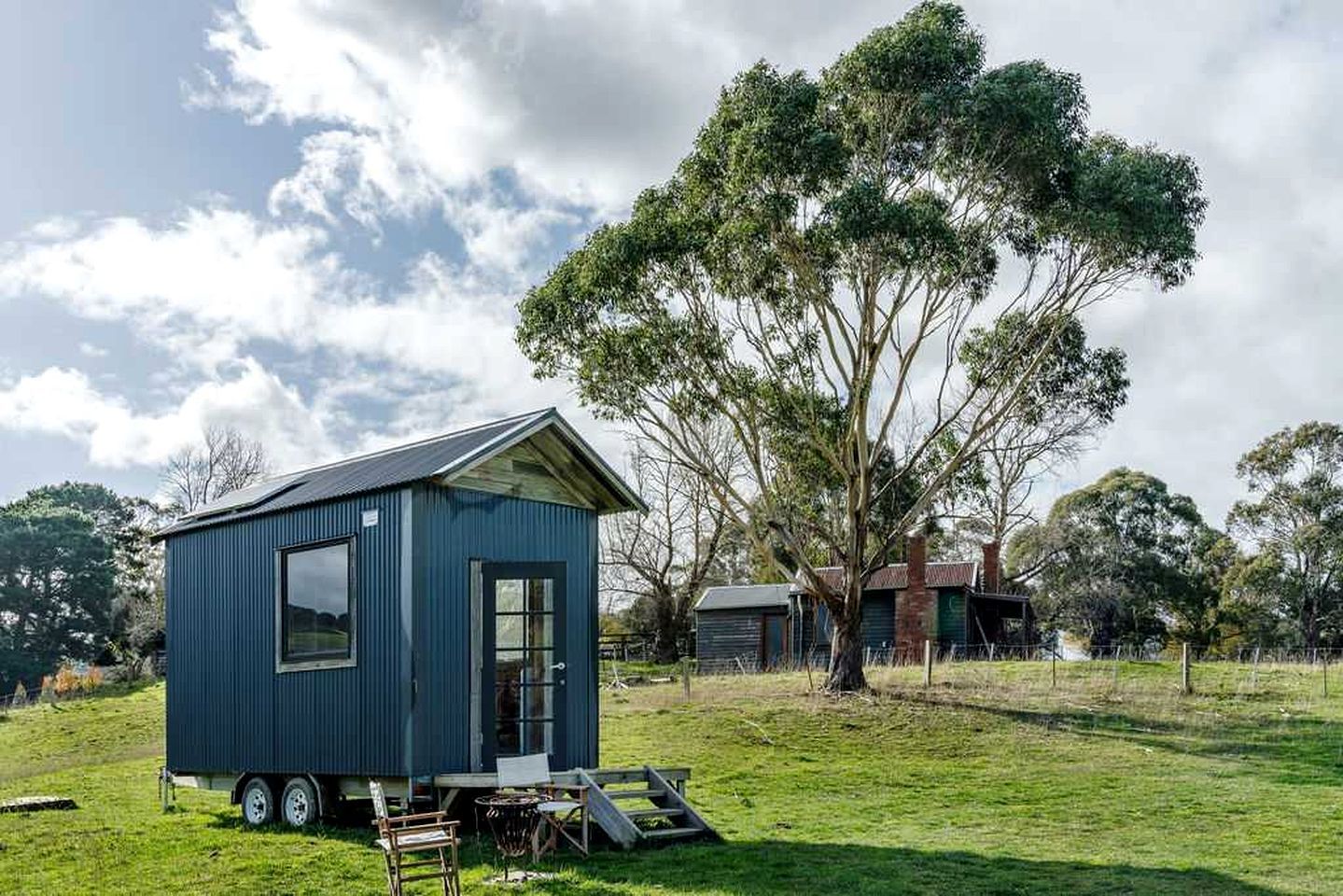Una pequeña casa móvil azul con remolque, una mesa de comedor y una silla en el exterior, rodeada de árboles y un campo cubierto de césped.