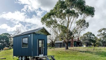Una pequeña casa móvil azul con remolque, una mesa de comedor y una silla en el exterior, rodeada de árboles y un campo cubierto de césped.