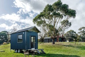 A small blue mobile home with a trailer, a dining table, and a chair outside, surrounded by trees and a grassy field.