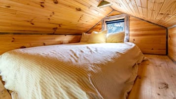 Une pièce chaleureuse, aménagée dans une cabane en bois, avec un plafond en pente, un lit simple recouvert d’une courtepointe blanche et une fenêtre laissant entrer la lumière naturelle.