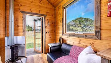 A wooden cabin interior with a fireplace, a couch, a table, and a large window offering a view of the outdoors.