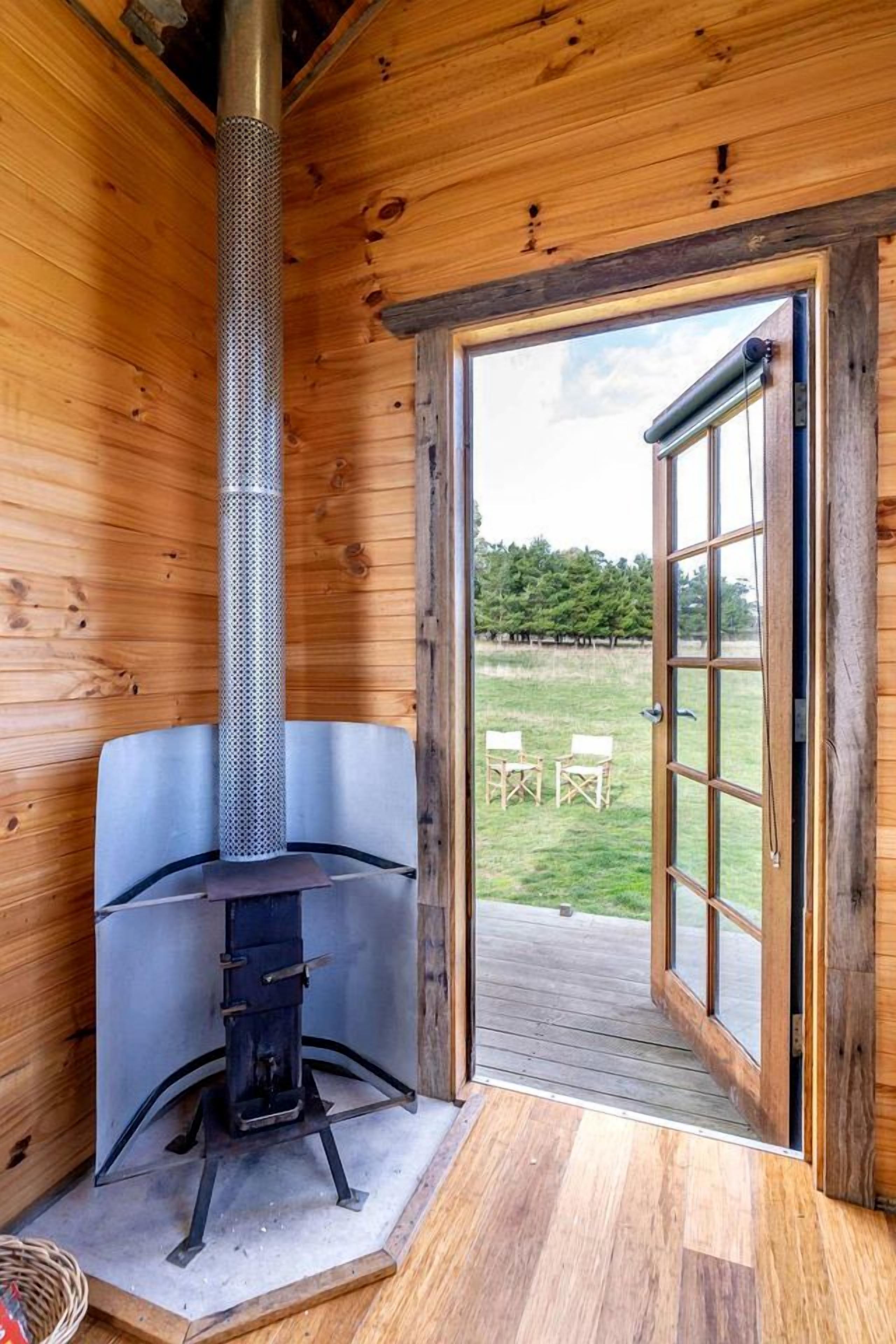Innenansicht einer Holzhütte mit einem Metallofen, einer Tür, die nach draußen führt, und Blick ins Freie.