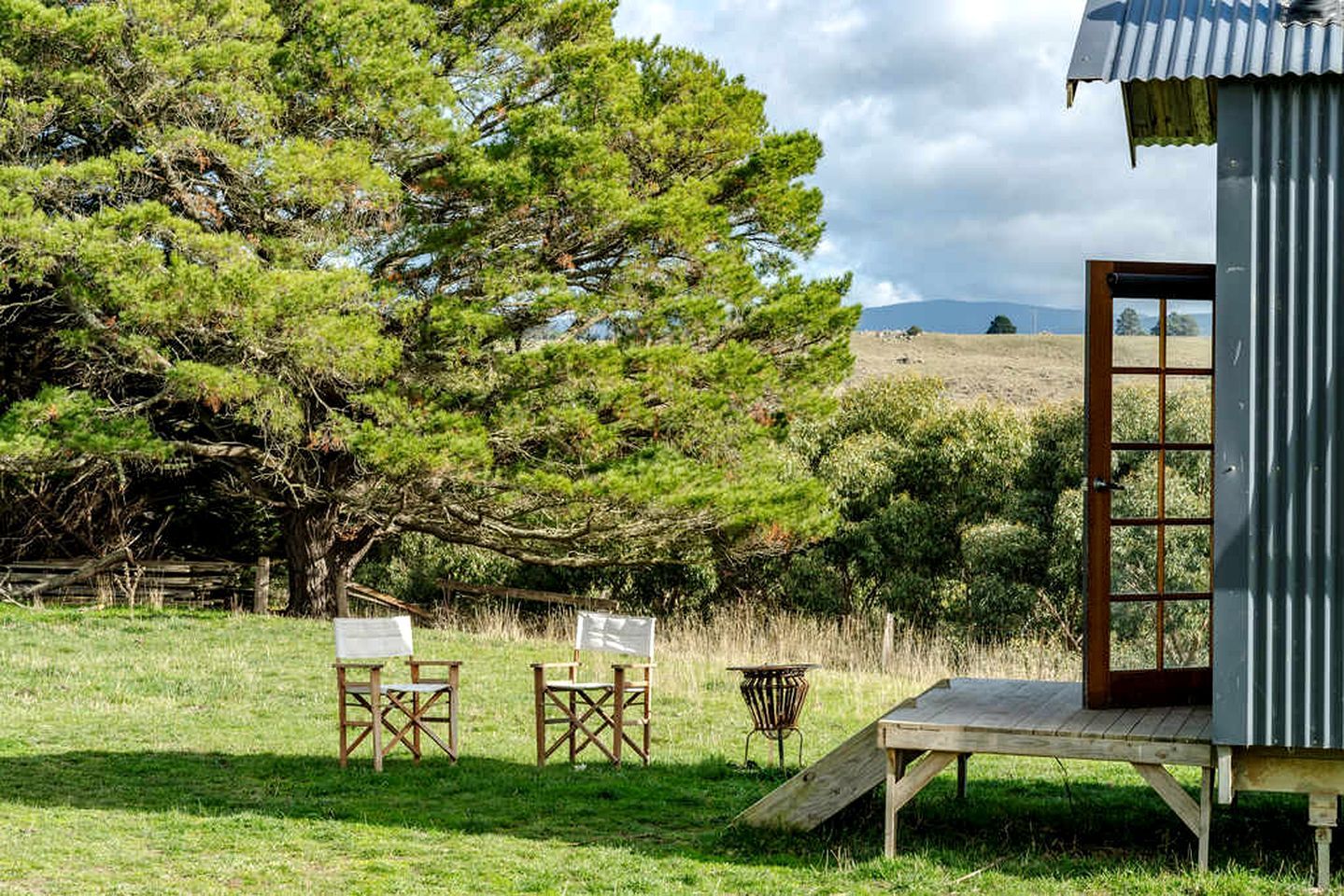 A rustic wooden structure with a ramp leading to a door, surrounded by a grassy field and trees.
