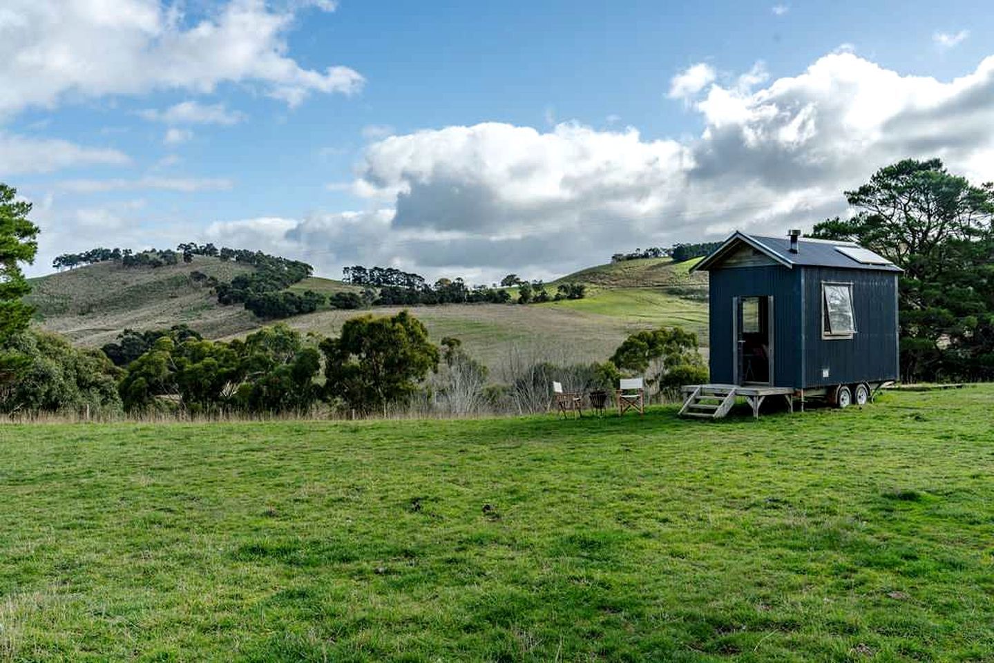 A small blue house with a porch, set in a grassy field with a hill in the background.