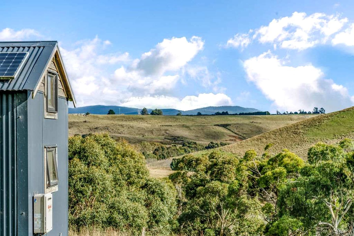 A house with a solar panel, surrounded by a hilly landscape with trees and a clear sky.