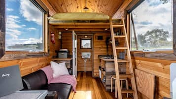 A compact interior with a wooden ceiling, a sofa, a ladder, and a window with a view of the outdoors.