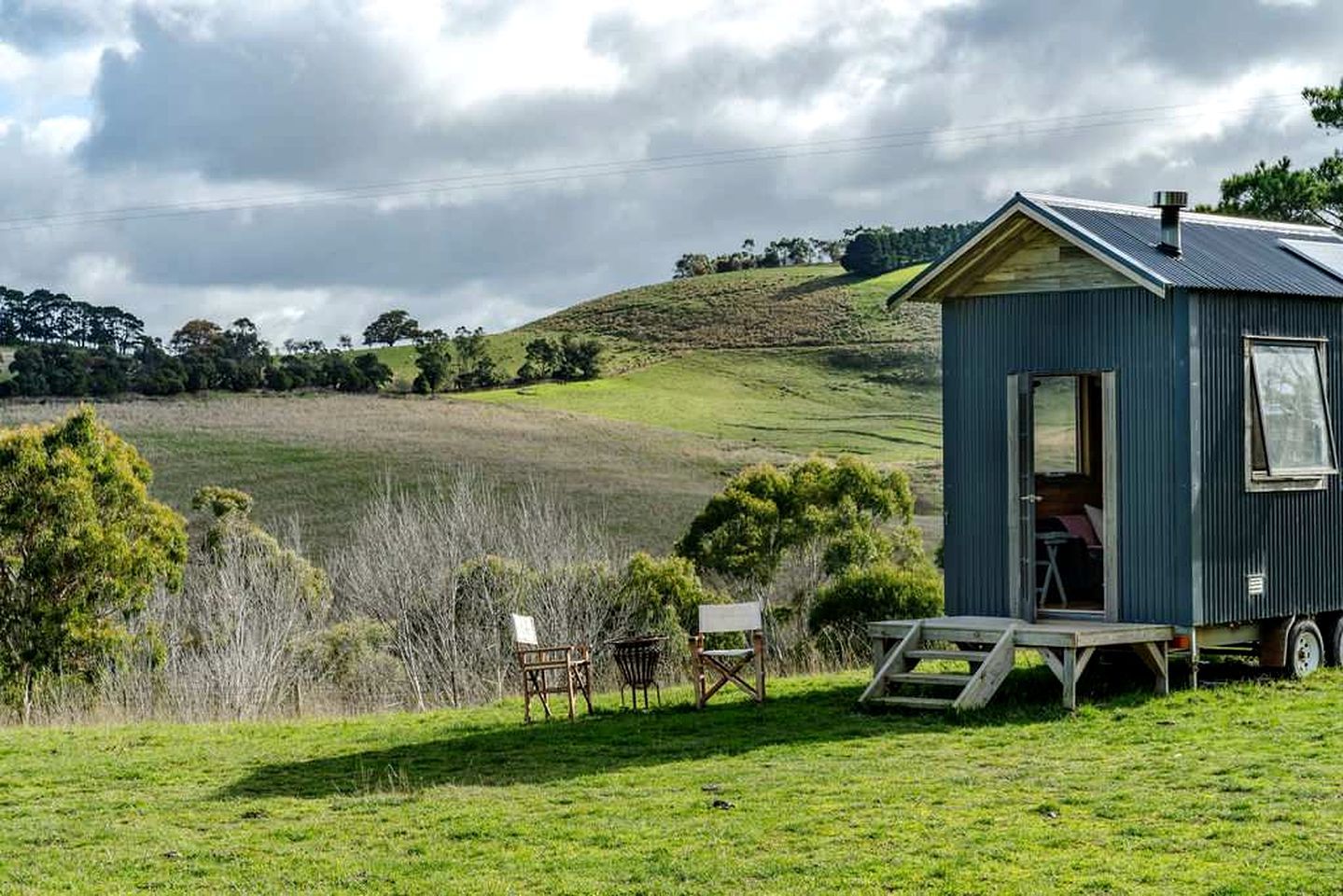 A small, single-story cabin with a metal roof, situated on a grassy field with a view of rolling hills in the background.