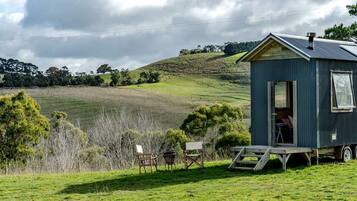 A small, single-story cabin with a metal roof, situated on a grassy field with a view of rolling hills in the background.