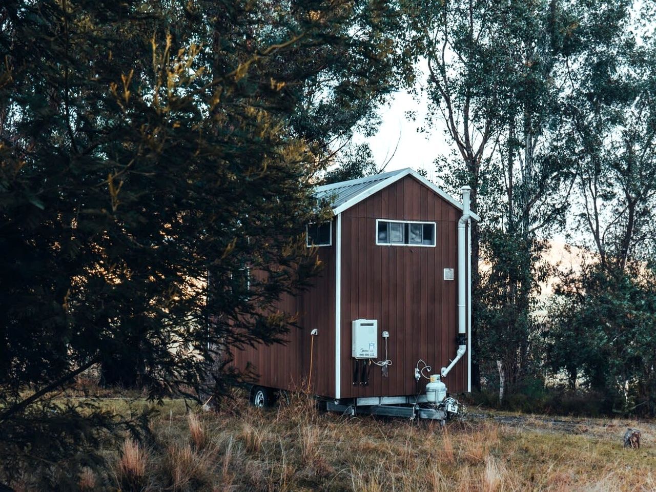 A small, single-story wooden structure with a sloped roof, surrounded by trees and grass.