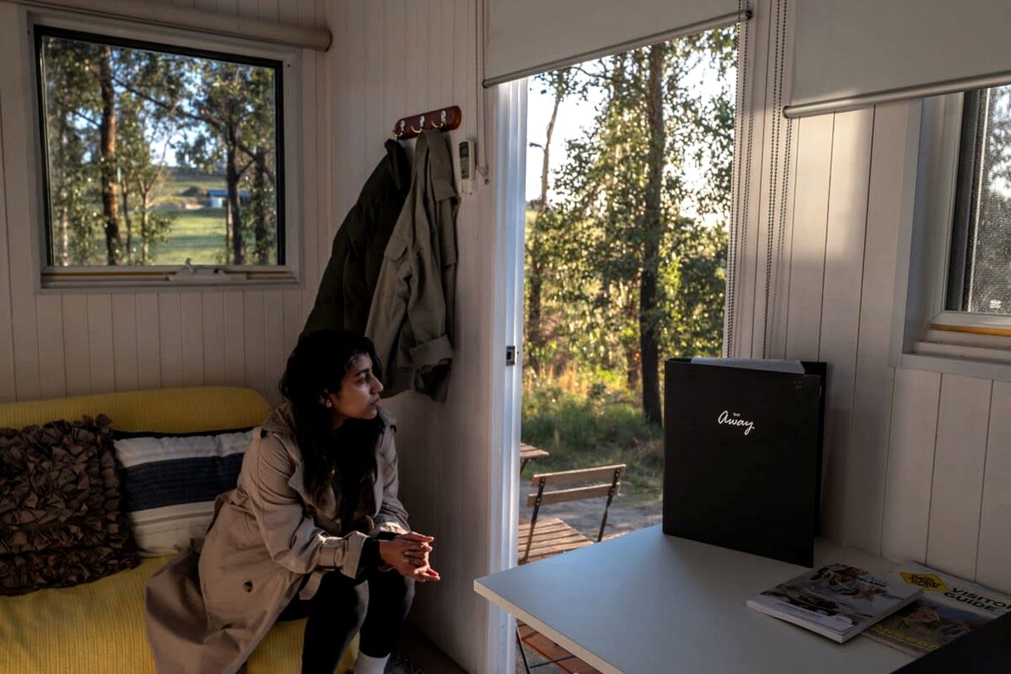 A room with a bed, a table with a magazine, a window with a view of trees, and a coat rack with jackets.