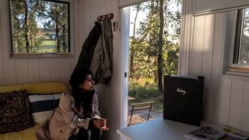A room with a bed, a table with a magazine, a window with a view of trees, and a coat rack with jackets.