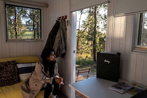A room with a bed, a table with a magazine, a window with a view of trees, and a coat rack with jackets.